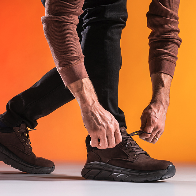 Men’s ProTech Verdell in Lince Dark Brown, shown on foot, with a man’s hands knotting the laces of the front shoe, on a gradient orange/yellow background